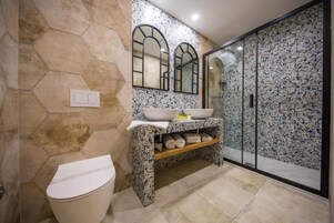 Uma casa de banho com parede revestida em azulejos hexagonais, sanita branca, lavatório com bancada de mármore e cabine de duche em vidro.