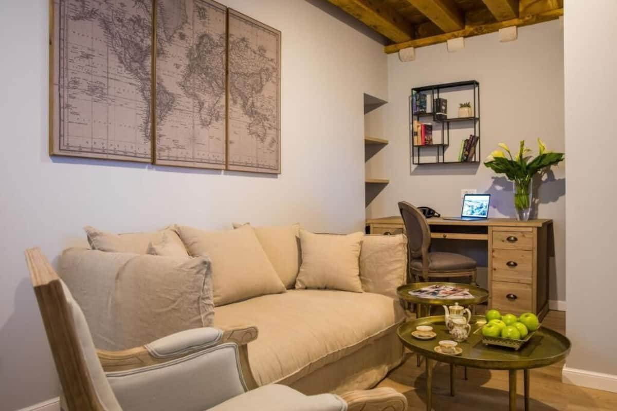 A cozy living room with a beige sofa, a wooden coffee table with a tray of green apples, a laptop on a desk, and a world map on the wall.