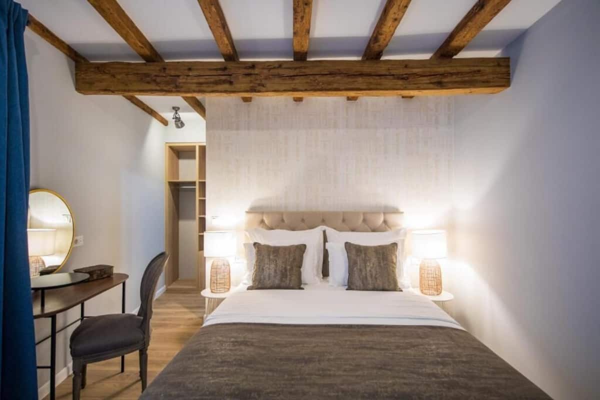 A bedroom with a large bed, two bedside lamps, a wooden ceiling, a desk with a chair, and a mirror.