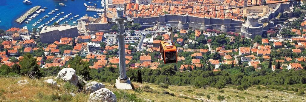 A cable car system with a cable car in motion, a historic fortification, and a coastal town with red-roofed buildings.