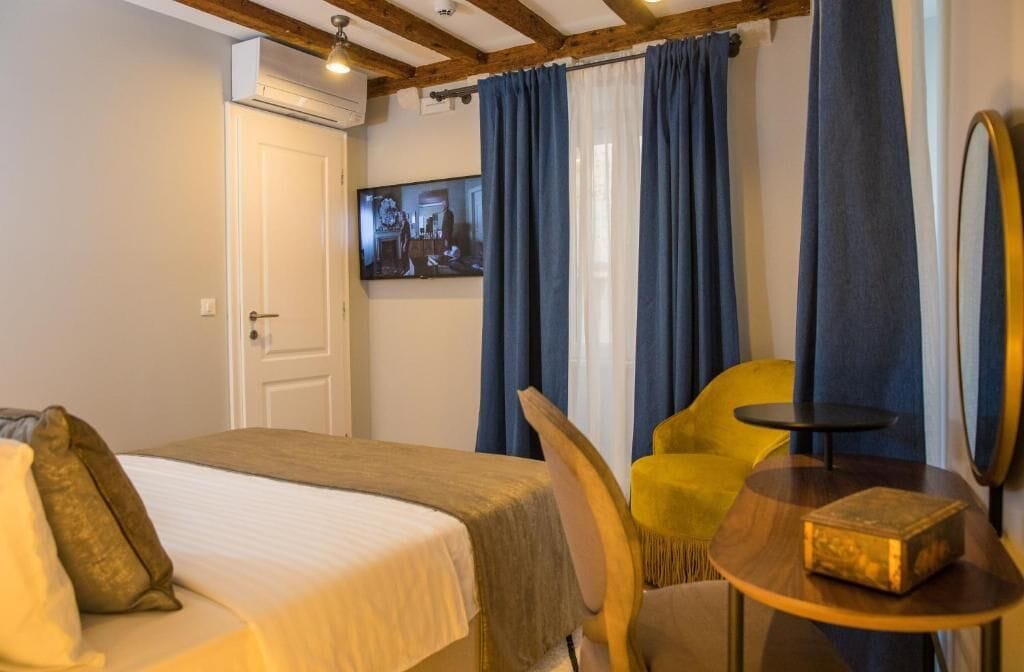 Habitación de hotel con cama, una silla amarilla, una mesita redonda y un televisor en la pared.