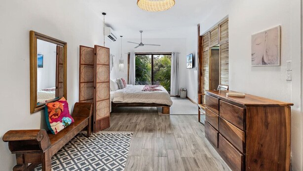 Een slaapkamer met een bed, een houten dressoir, een spiegel, een plafondventilator en uitzicht op groen buiten.