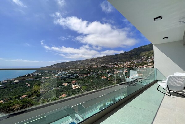 Un balcon doté d’une balustrade en verre, offrant une vue sur une ville côtière et la mer.