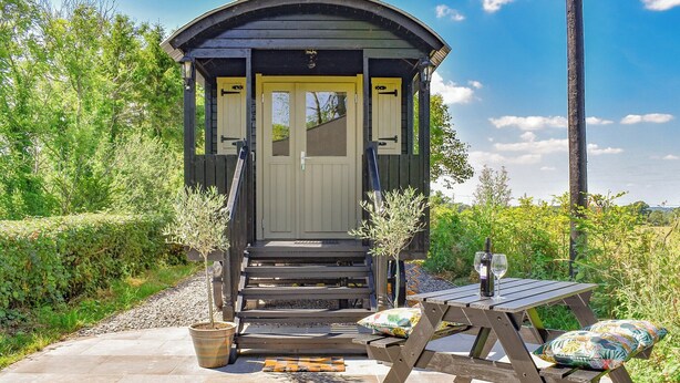 Eine kleine Holzhütte mit Tür und Stufen, umgeben von Grünpflanzen und einem Picknicktisch mit Kissen.