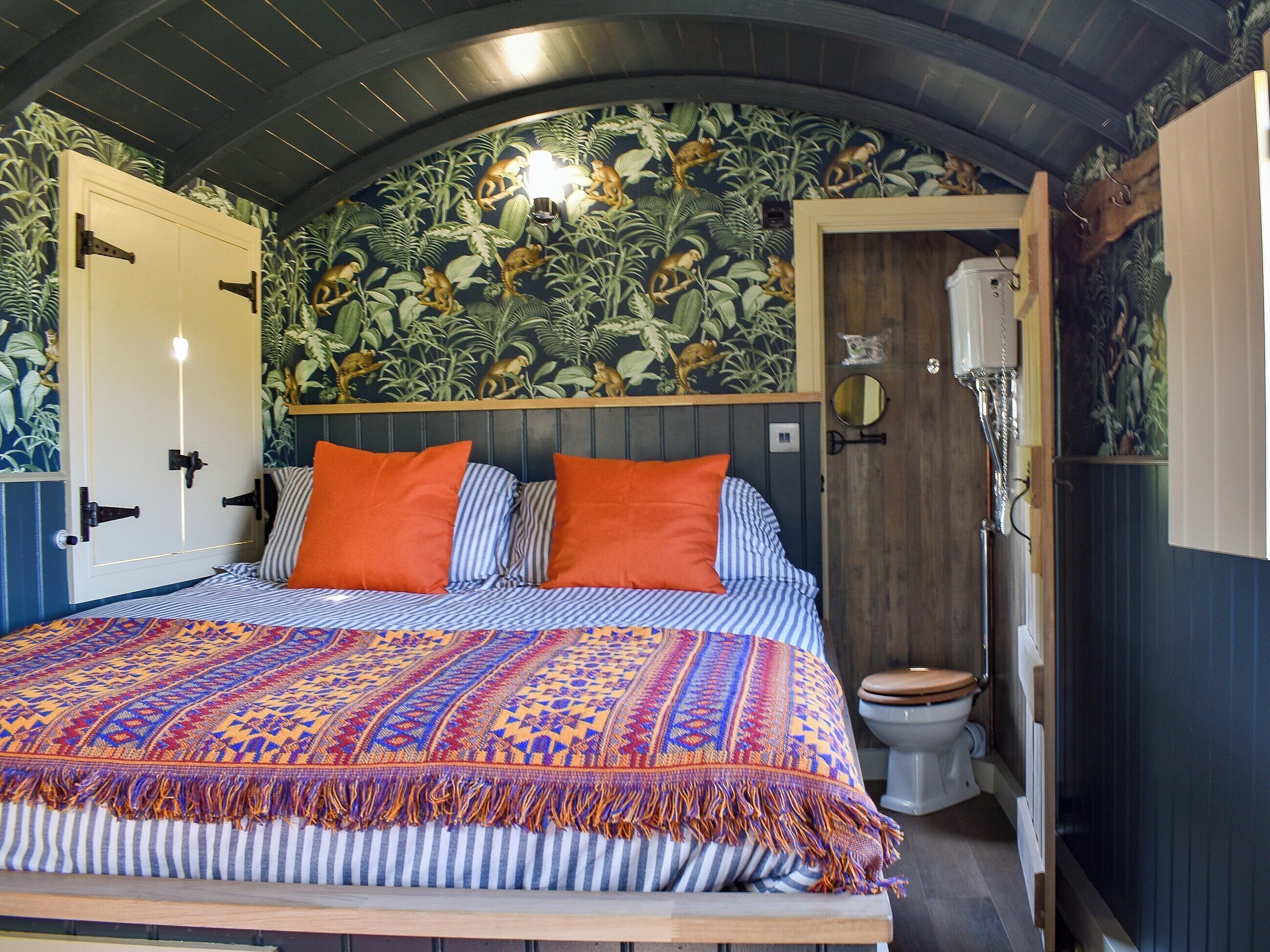 A bedroom with a bed, two orange pillows, a patterned blanket, a wooden headboard, a wall with a monkey mural, and a wooden door.