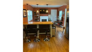 A kitchen with a wooden bar, four bar stools, pendant lighting, a white fireplace, and a refrigerator.