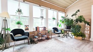 A sunlit room with large windows, a black chair, a wooden table, and various plants. - Boho, plant-filled home w/ wood + leather touches, 3 beds, 2 baths, super clean. (Emerald Isle)