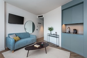 A modern living room with a sofa, a coffee table, a TV, and a kitchen area with cabinets and a coffee machine.