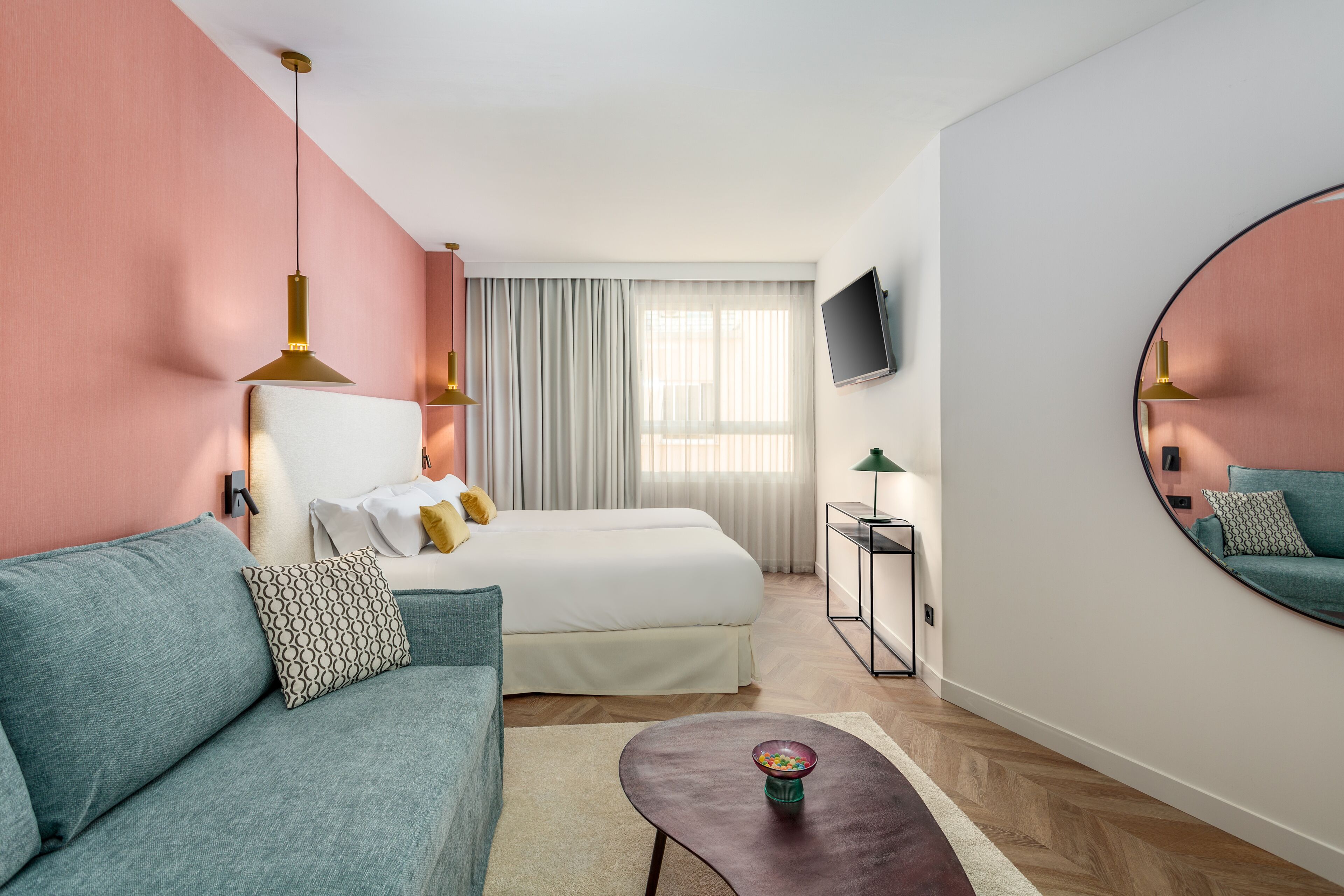 A modern hotel room with a bed, a sofa, a small table, a TV, and a mirror.