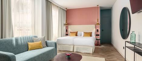 A modern hotel room with a teal sofa, a wooden coffee table, a double bed with white bedding, and a large mirror on the wall.