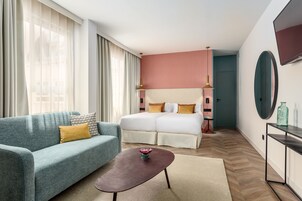 A modern hotel room with a teal sofa, a wooden coffee table, a double bed with white bedding, and a large mirror on the wall.