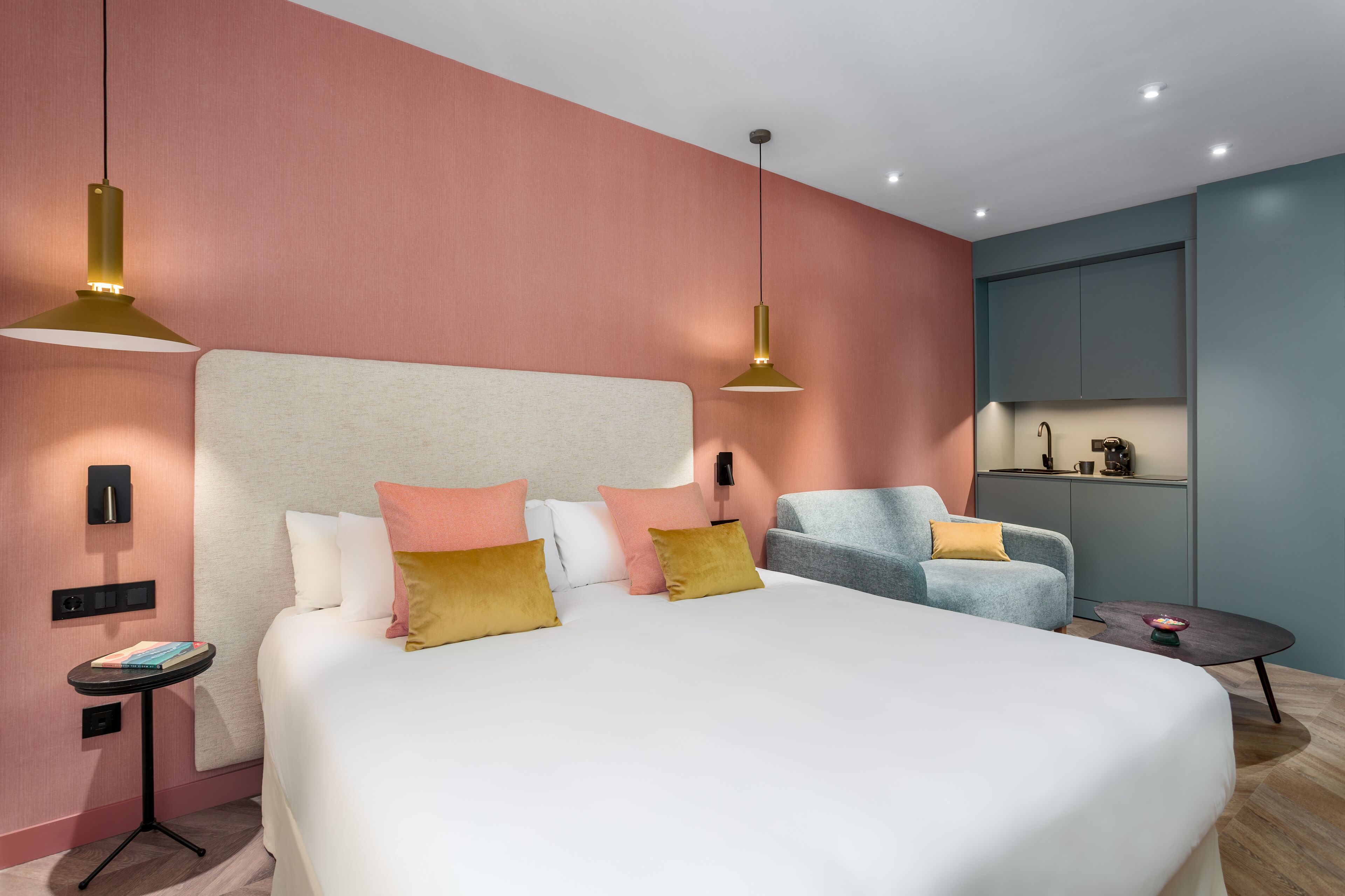 A modern hotel room with a large bed, a grey armchair, a small round table, and a kitchenette area.