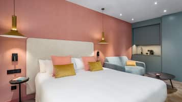 Una habitación de hotel moderna con una cama grande, un sillón gris, una mesita redonda y una zona de kitchenette.