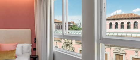 Habitación de hotel con un ventanal grande, una cama con almohadas blancas y melocotón, y vistas a edificios históricos.