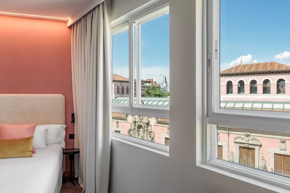 A hotel room with a large window, a bed with white and peach pillows, and a view of historic buildings. - Tribu Malasaña (Madrid)