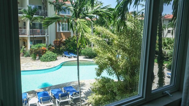 Vista de una piscina con sillas de descanso y palmeras, rodeada por un edificio de varios pisos con balcones.