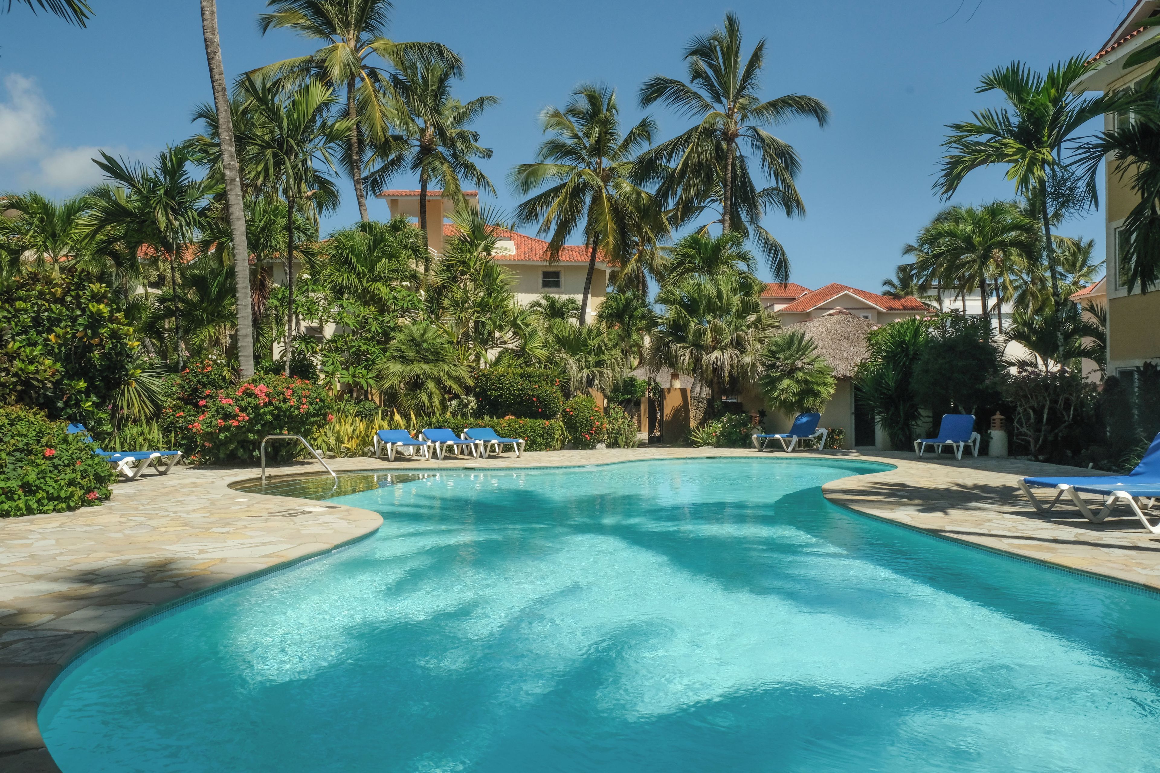 A swimming pool with lounge chairs, surrounded by palm trees and a building with a red-tiled roof.