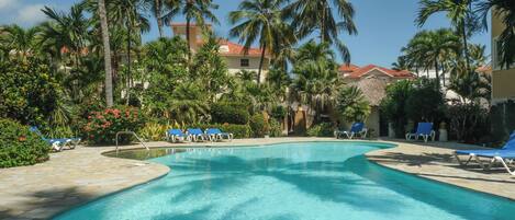 A swimming pool with lounge chairs, surrounded by palm trees and a building with a red-tiled roof.