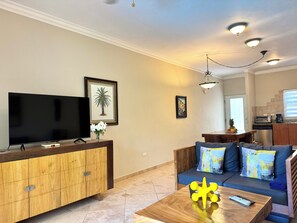 A living room with a flat-screen TV, wooden cabinet, sofa with cushions, and a coffee table with a remote.