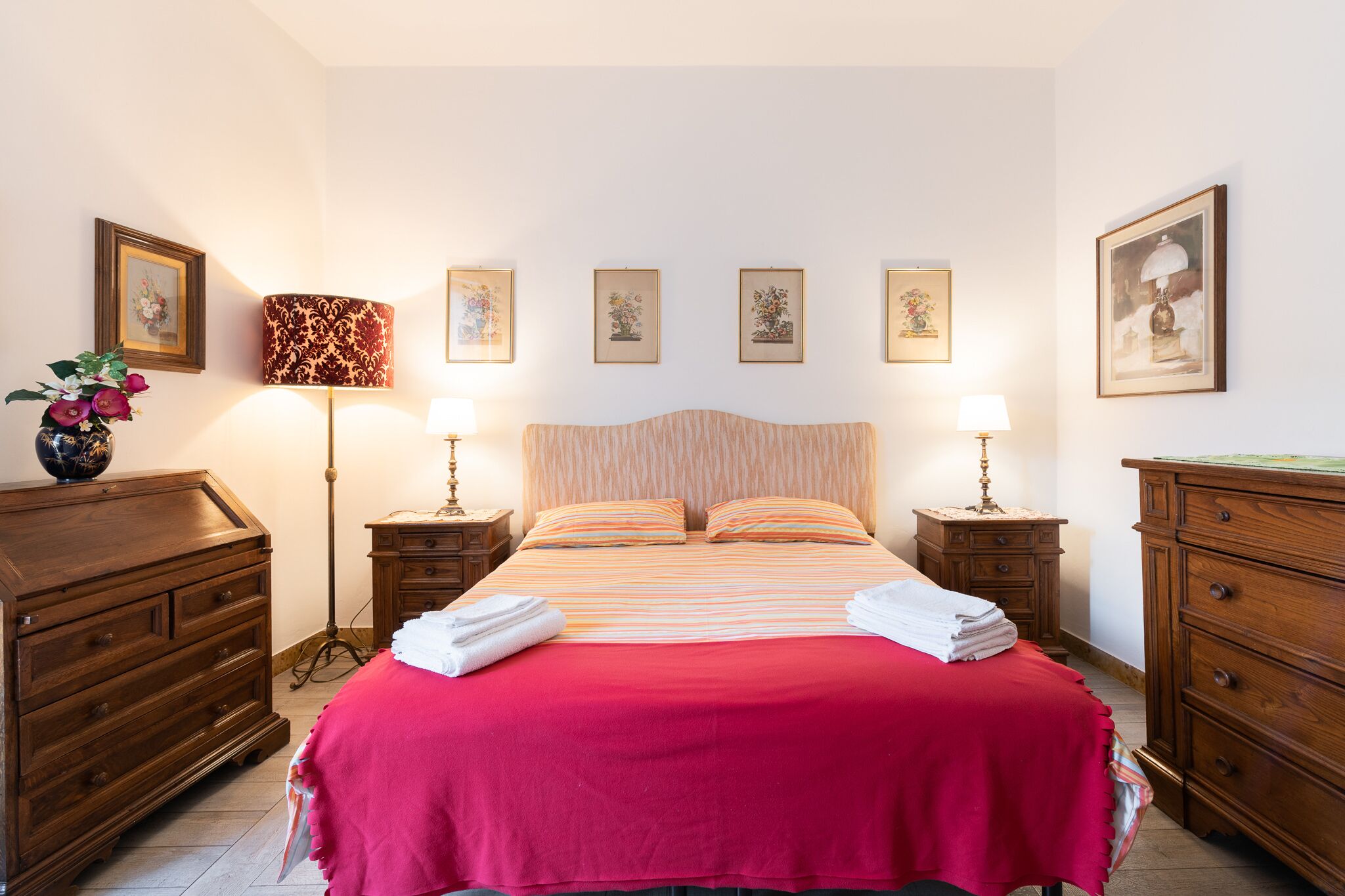 Cama bem arrumada com colcha vermelha, acompanhada por duas mesas de cabeceira com luminárias, um aparador de madeira e obras de arte emolduradas nas paredes.