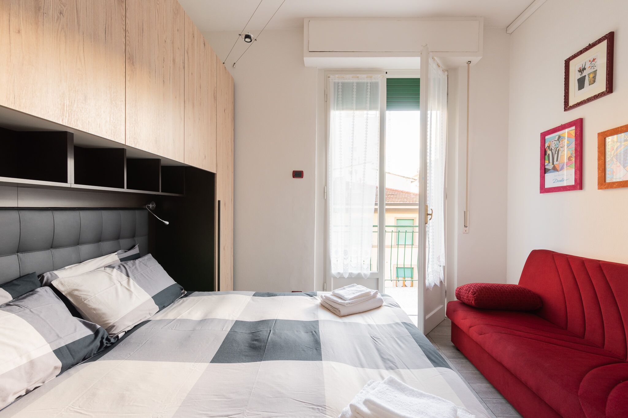 Un dormitorio con cama, sofá rojo, armarios de madera y un balcón con vistas.