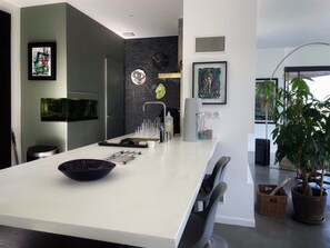 A modern kitchen with a white countertop, black chairs, and a large island. There are framed artworks on the walls and a potted plant on the right.