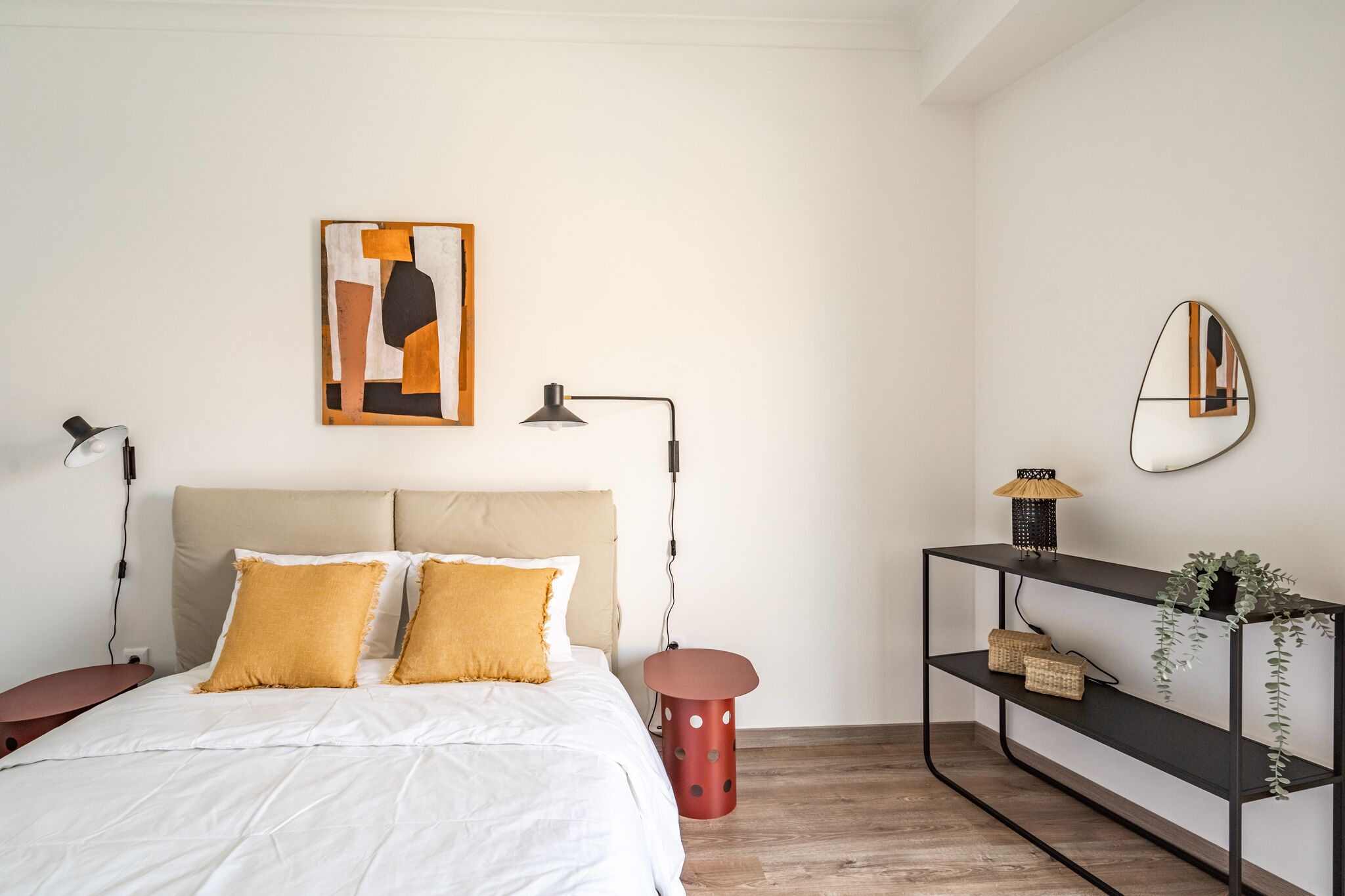 A bedroom with a bed, a bedside table, a lamp, a wall-mounted artwork, a mirror, and a console table with shelves.