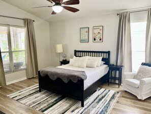 Un dormitorio con una cama, una mesita de noche, una silla, un ventilador de techo y dos cuadros en la pared.