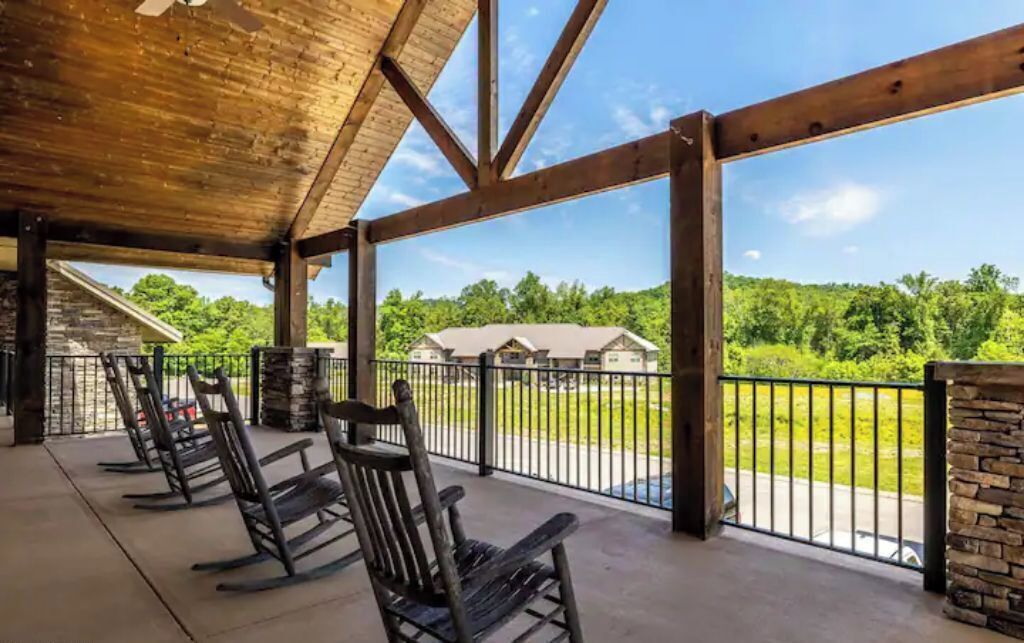 A covered outdoor seating area with wooden rocking chairs, stone pillars, and a view of a green landscape.