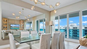 A modern hotel room with a glass dining table, white chairs, a large window with a city view, and a chandelier. - Premium 3-Bedroom Oceanview Penthouse at 1 Hotel (Miami Beach)