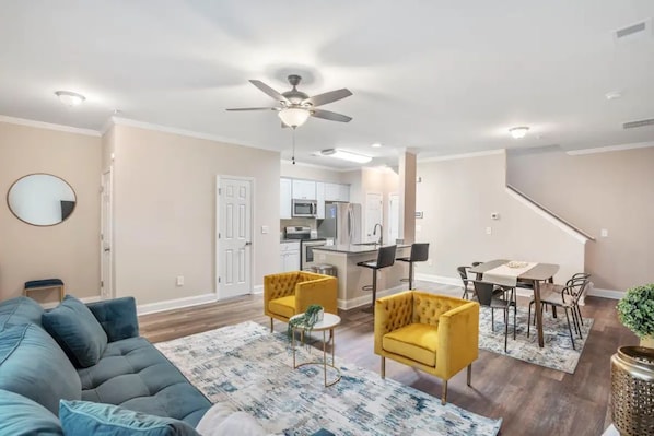 Un salon moderne avec un canapé, deux fauteuils, une table à manger avec des chaises, une cuisine en arrière-plan et un ventilateur de plafond.