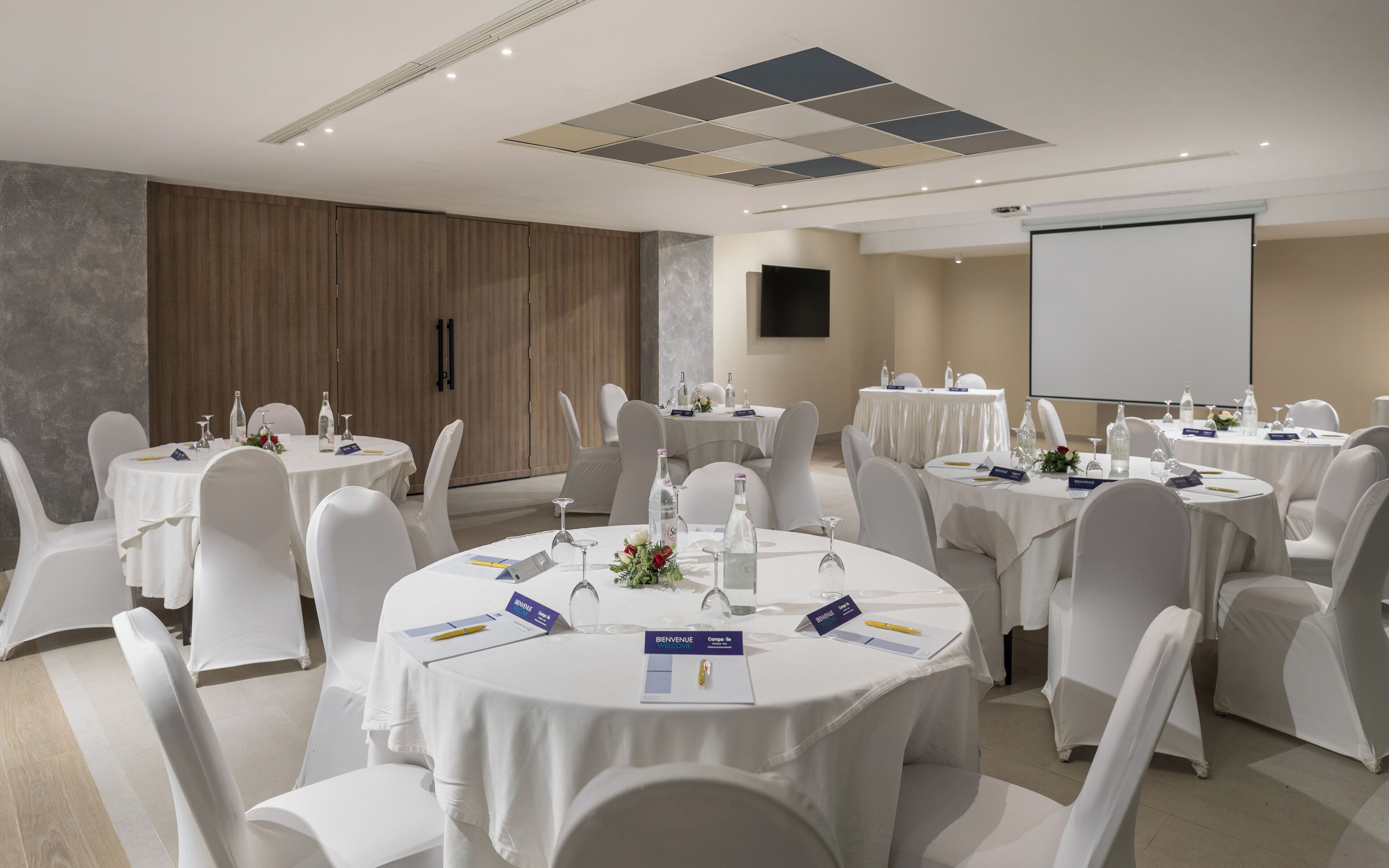 A conference room with round tables, white chairs, nameplates, and a projector screen.