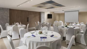 Une salle de conférence équipée de tables rondes, de chaises blanches, de plaques nominatives et d’un écran de projection.
