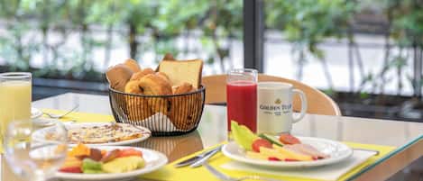 Una mesa de desayuno con cruasanes, tostadas, una bandeja de fruta, un vaso de jugo de naranja y un vaso de jugo rojo.