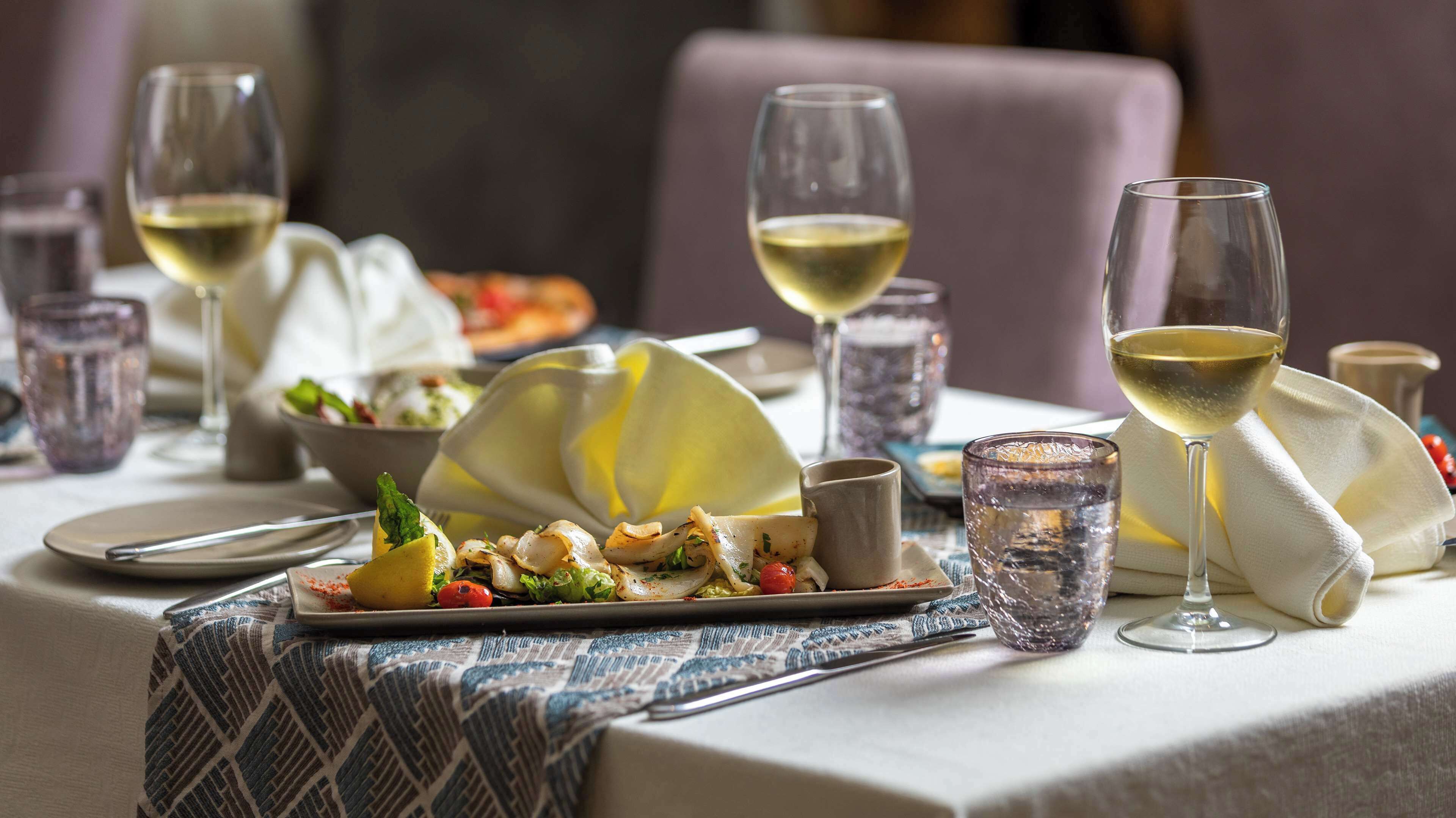 A dining table set with plates of food, glasses of white wine, and a napkin.