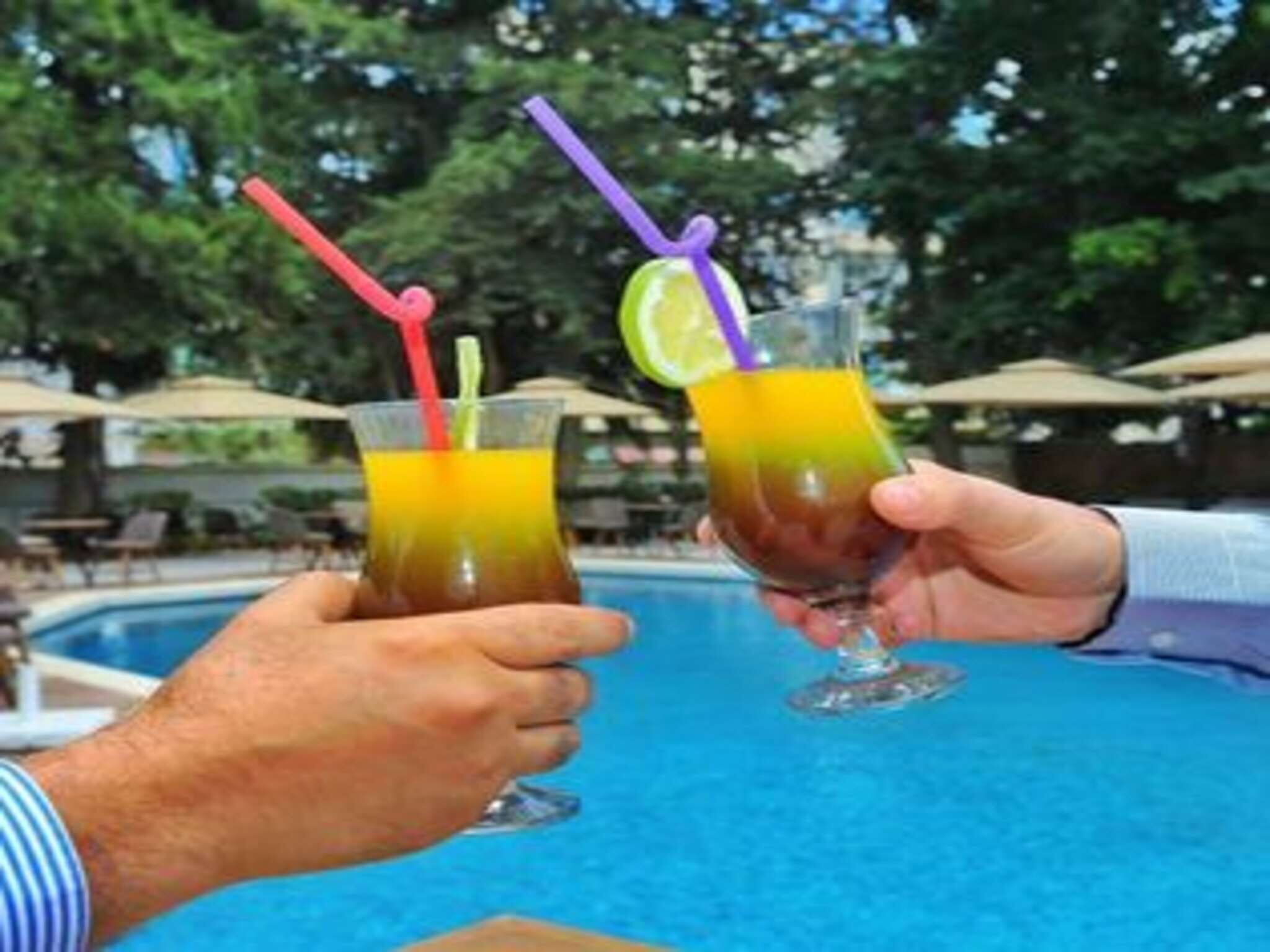 Two people holding glasses of drinks with straws and lime slices by a pool.