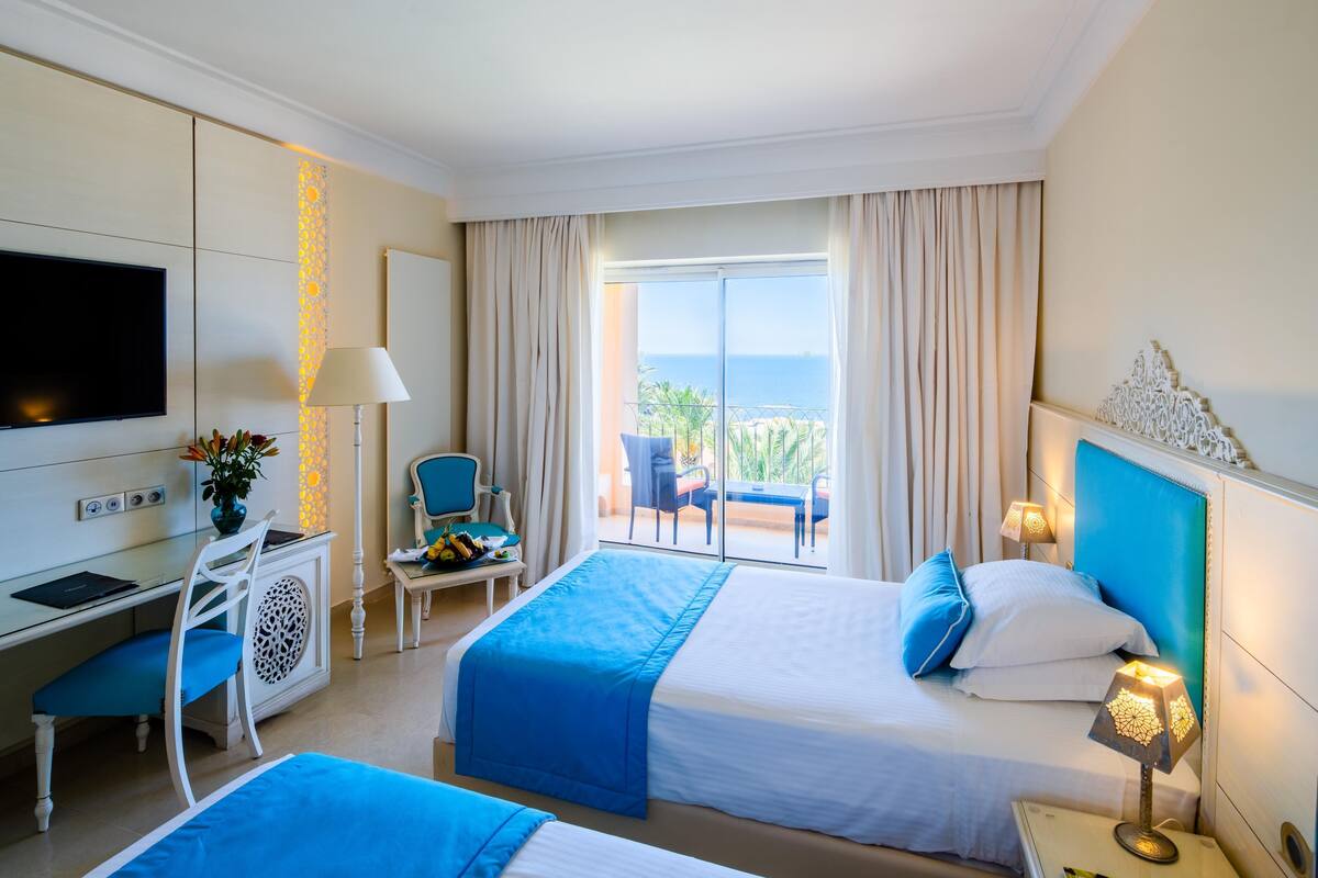 A hotel room with two beds, a desk, a chair, a TV, and a balcony with a view of the sea.