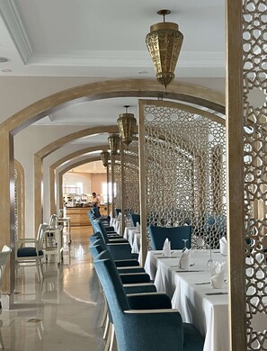 A restaurant with a long dining area, white tables, blue chairs, and decorative hanging lamps. - Royal Tulip Korbous Bay Thalasso & Springs (Qurbus)