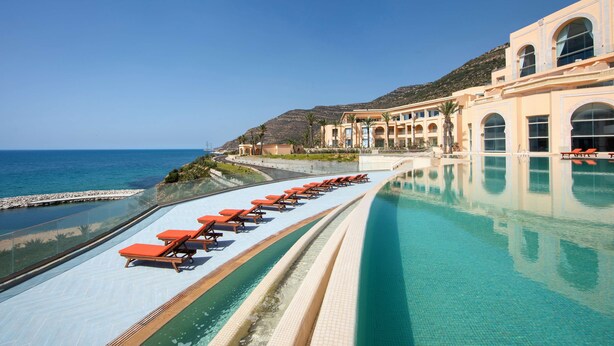 A poolside view with lounge chairs, a clear blue sky, and a building with arched windows and balconies overlooking the sea. - Royal Tulip Korbous Bay Thalasso & Springs (Qurbus)