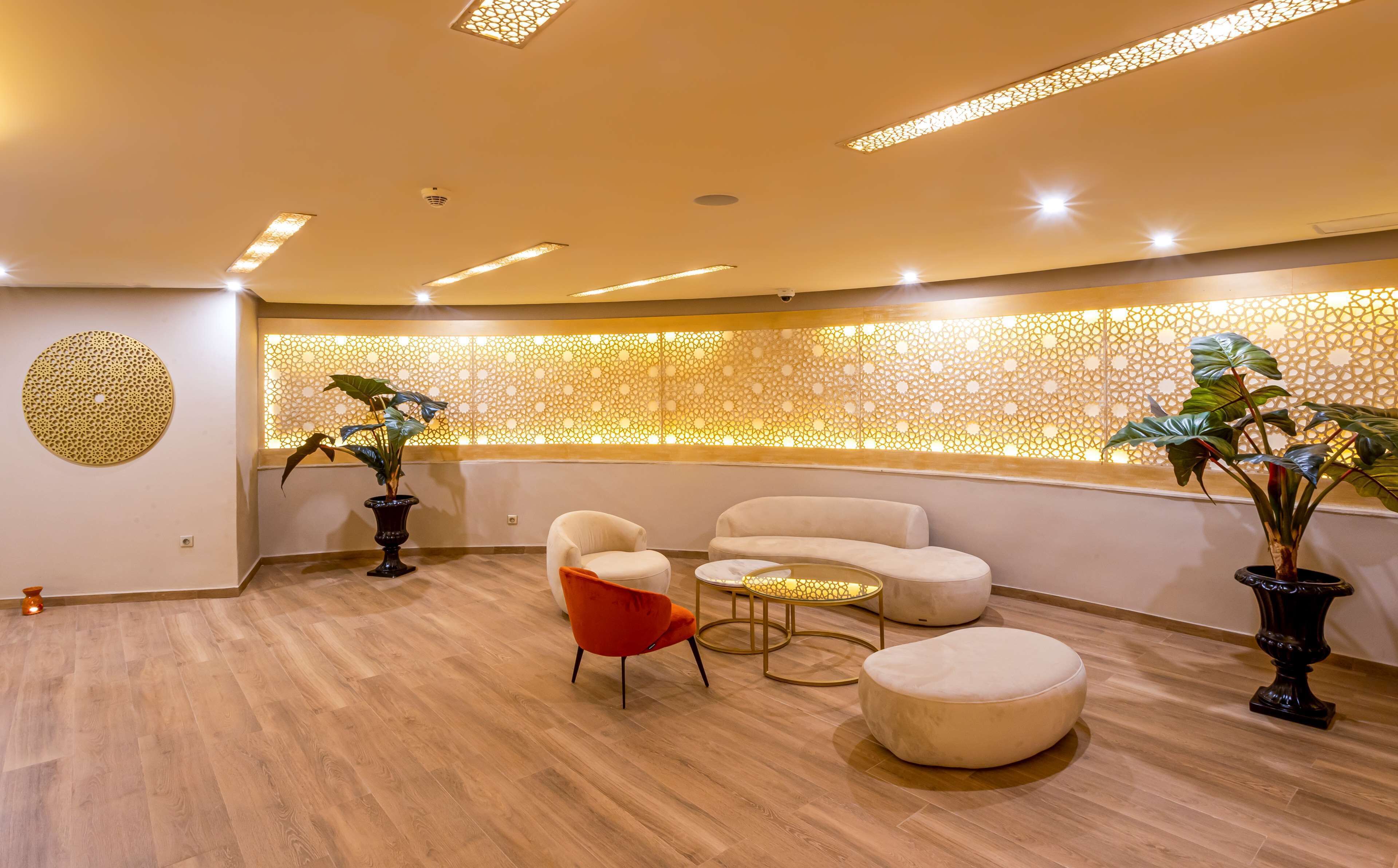 A modern lounge area with a wooden floor, a sofa, a red chair, a small table, and decorative plants.