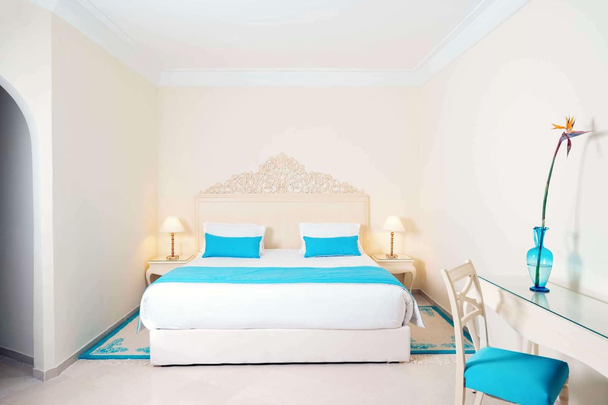 A hotel room with a large bed, two bedside tables with lamps, a glass table with a vase, and a chair.