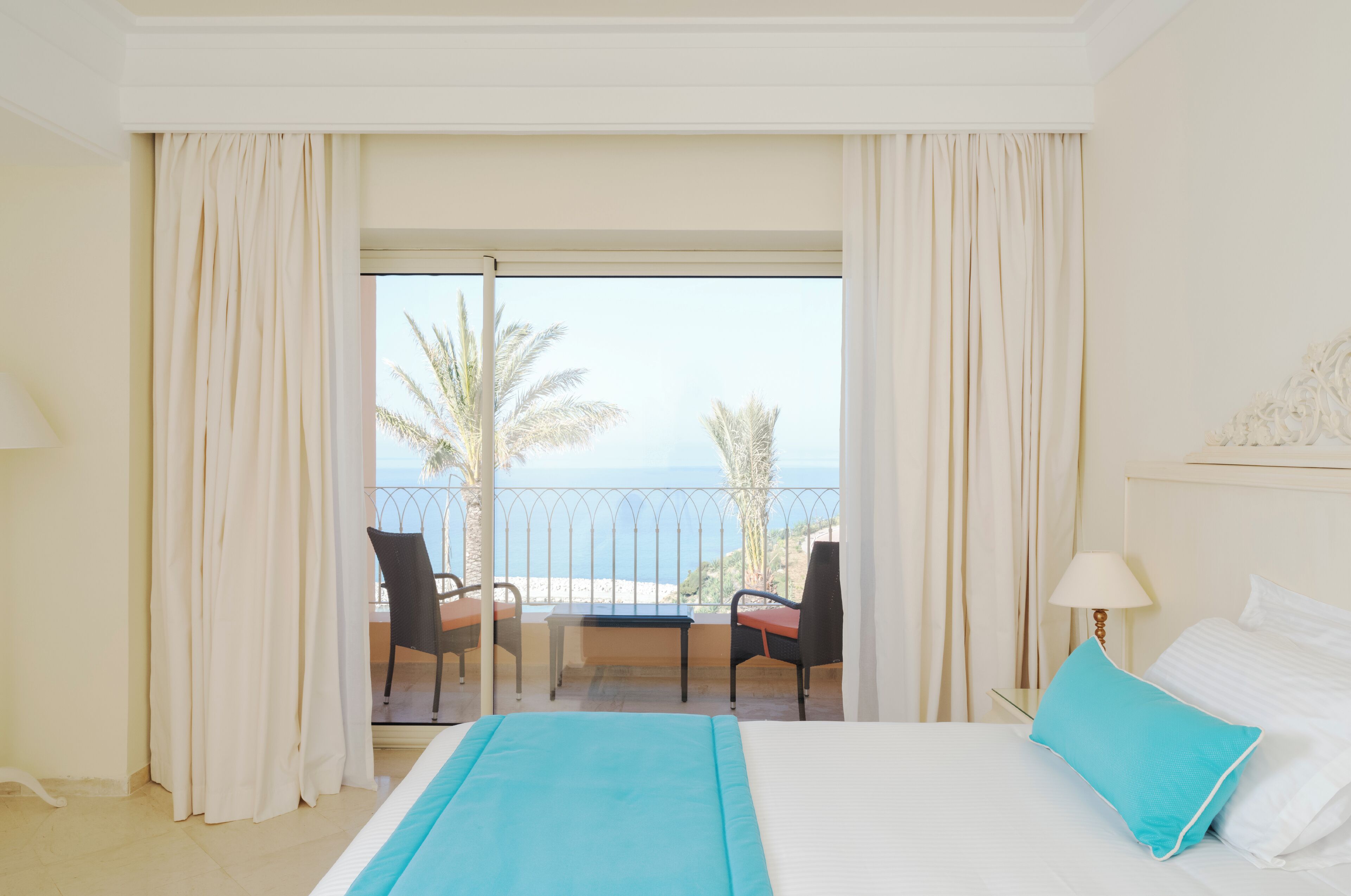 Habitación de hotel con balc ón, cama con ropa de cama blanca y azul, mesita de noche con lámpara y vistas a la playa y palmeras.