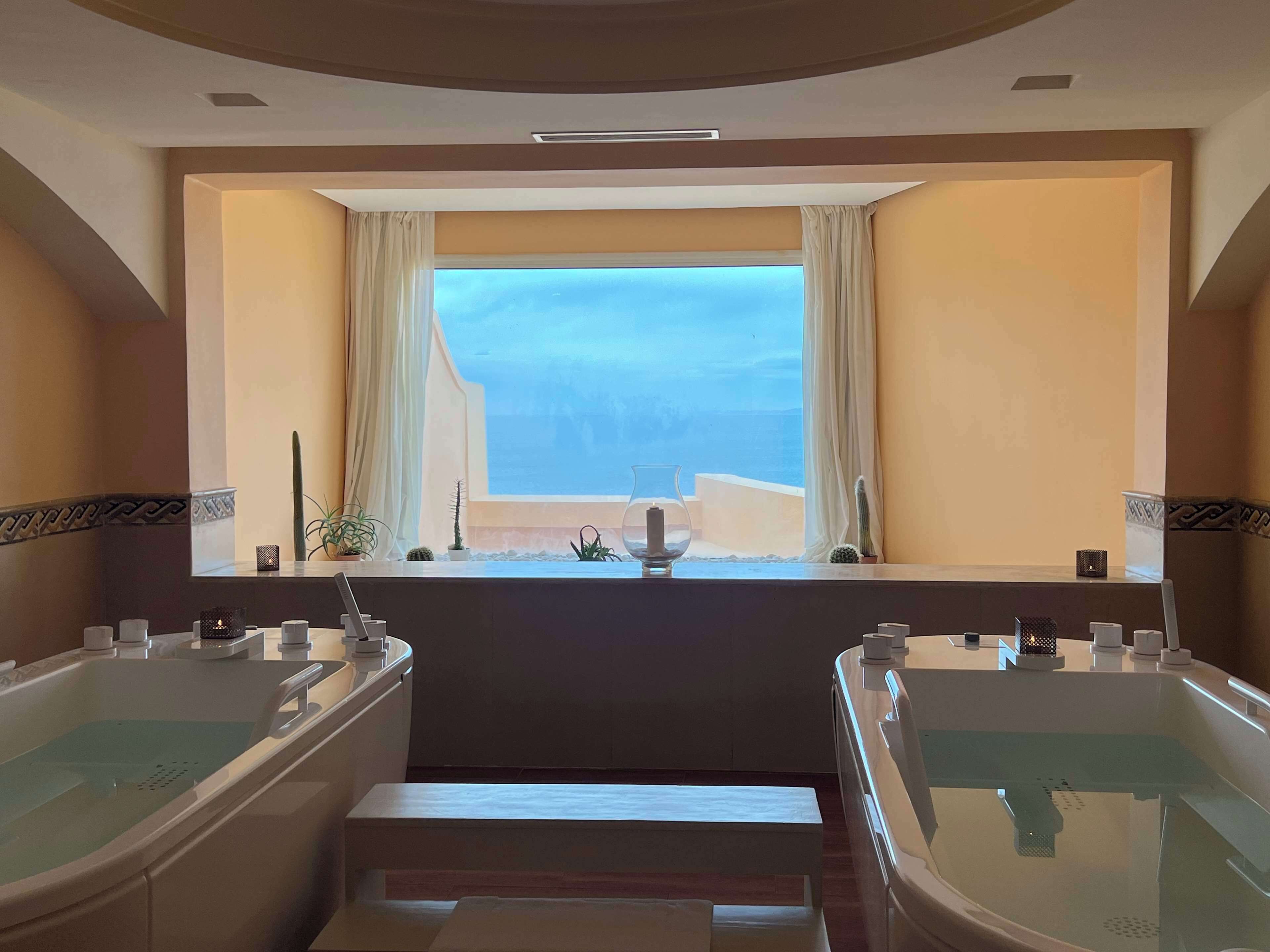Un baño con dos bañeras grandes, una ventana con cortinas y vistas al mar.