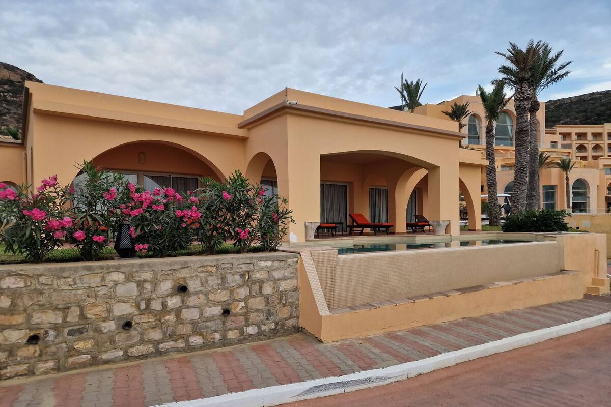 A hotel with a pool, stone wall, and red lounge chairs.