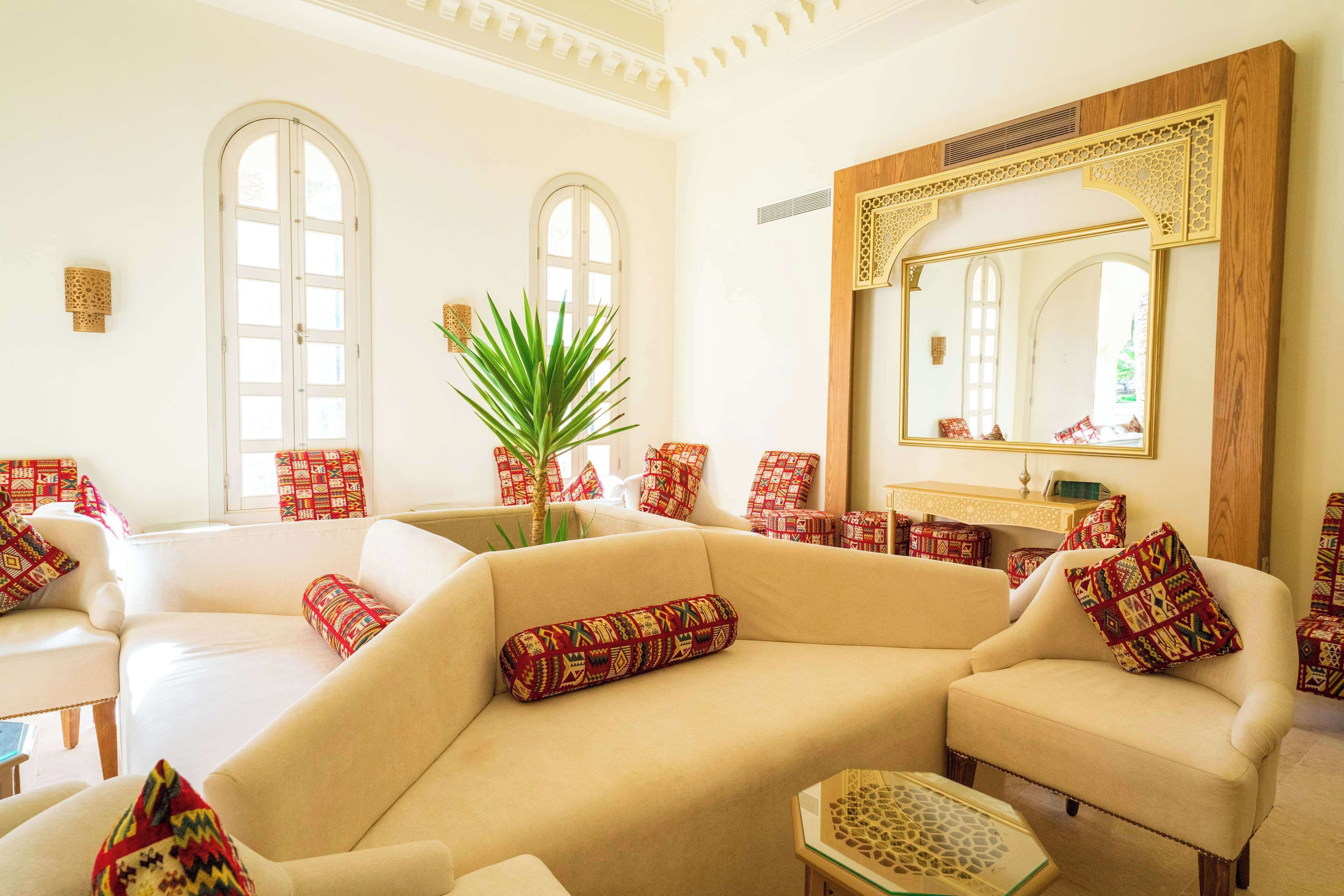 A spacious living room with a beige sofa, patterned cushions, a large mirror, and a potted plant.
