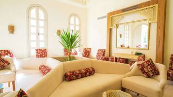 A spacious living room with a beige sofa, patterned cushions, a large mirror, and a potted plant.