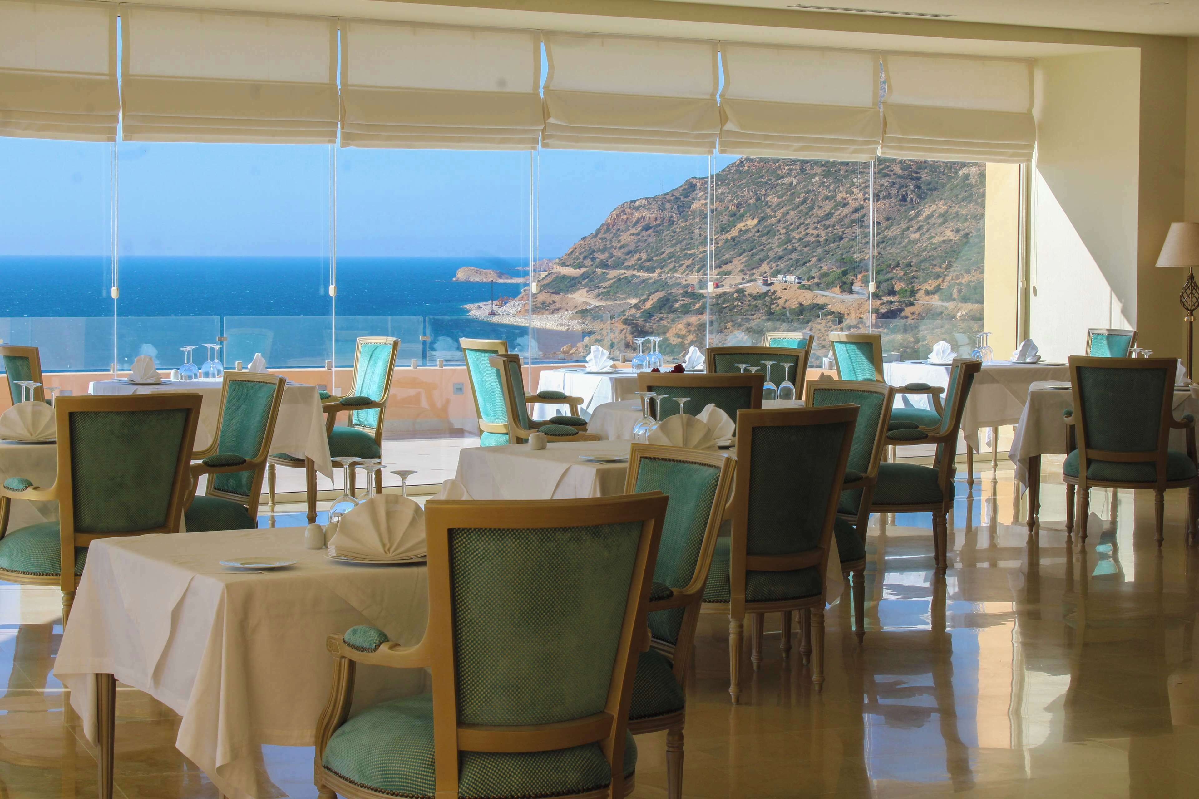A restaurant with tables set for dining, chairs with green cushions, and a view of the sea and a hillside.