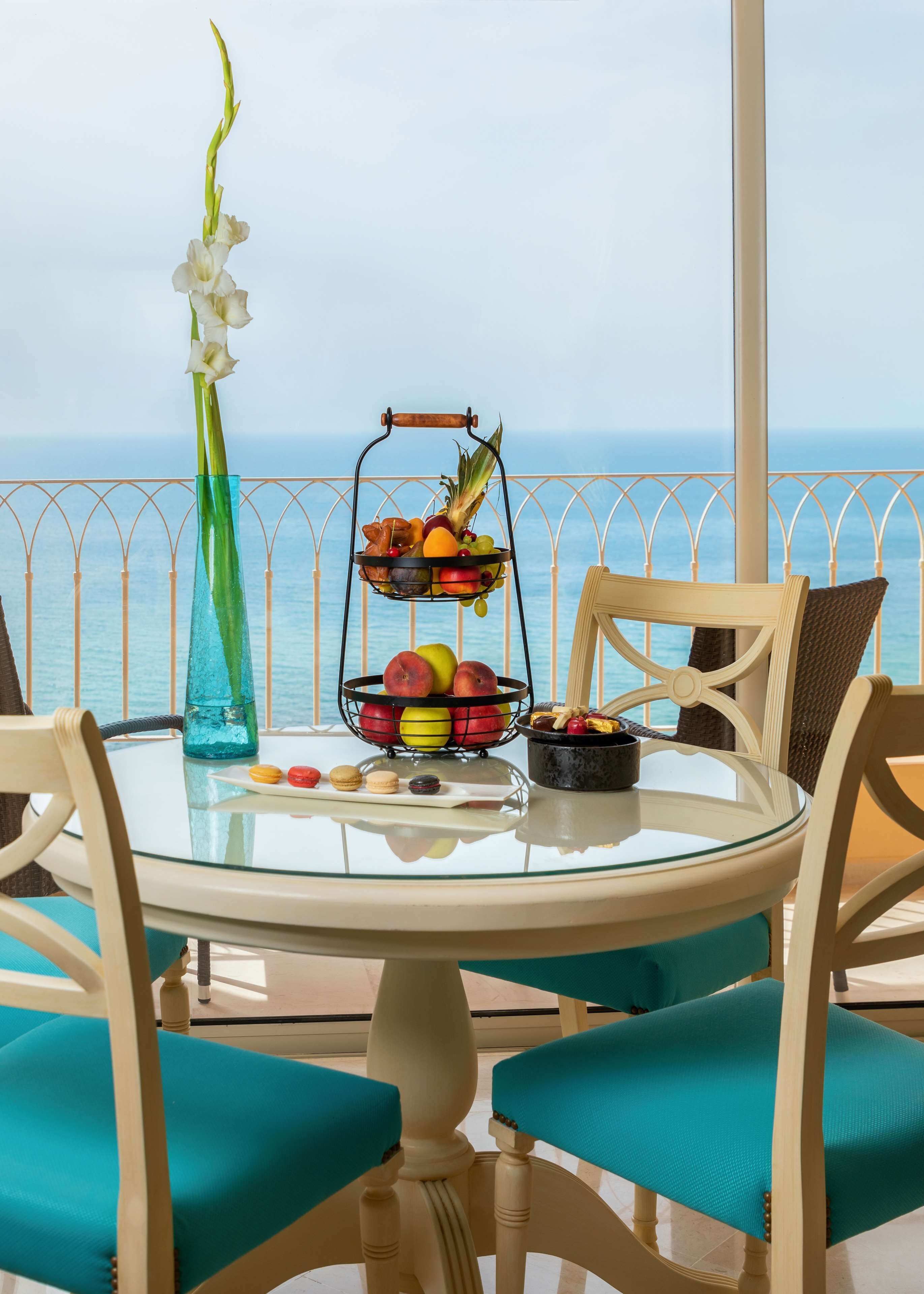 Una mesa redonda de vidrio con una cesta de frutas, un jarrón con una flor y un pequeño recipiente con macarons en un balcón con vista al mar.