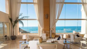 A spacious living room with a large window offering a view of the ocean, a white sofa, a glass coffee table, and a potted plant. - Royal Tulip Korbous Bay Thalasso & Springs (Qurbus)