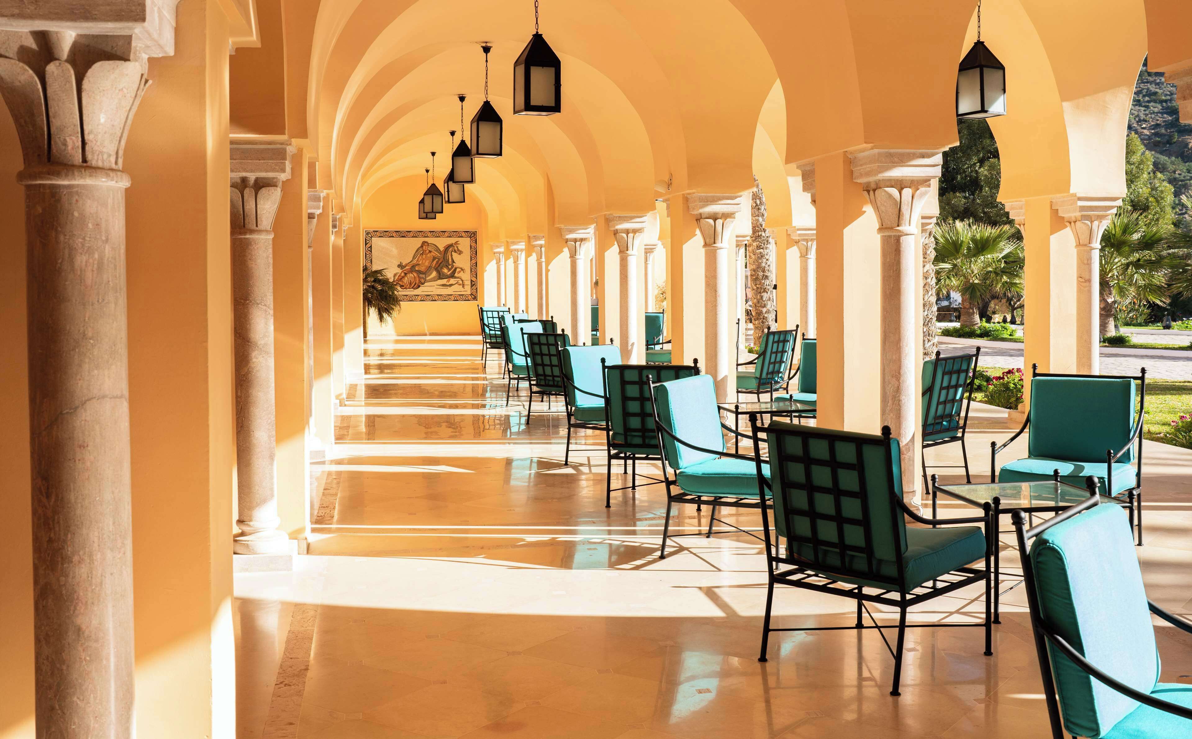 A covered outdoor seating area with rows of chairs and tables, supported by columns, and featuring hanging lanterns.